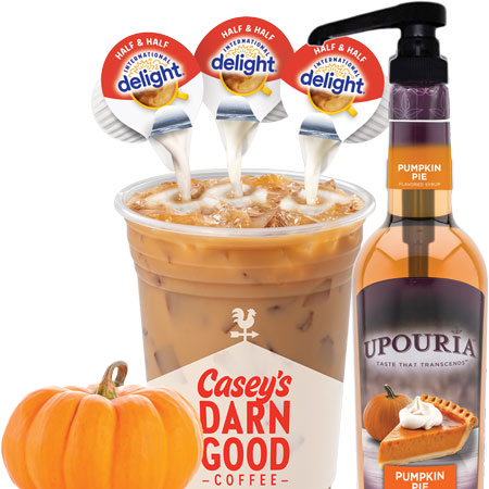 Cup of iced coffee with half and half being poured in next to a bottle of pumpkin syrup adorned with a tiny pumpkin.