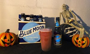 A 6 pack of Blue Moon in front of a Halloween background with a Spooky Mist cocktail